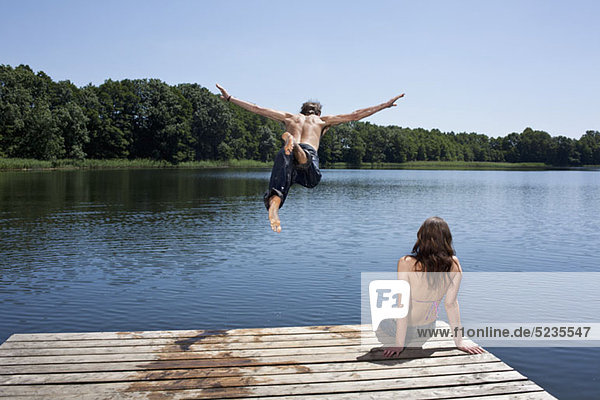 Der Typ taucht mit ausgestreckten Armen in den See  während das Mädchen am Steg zuschaut.