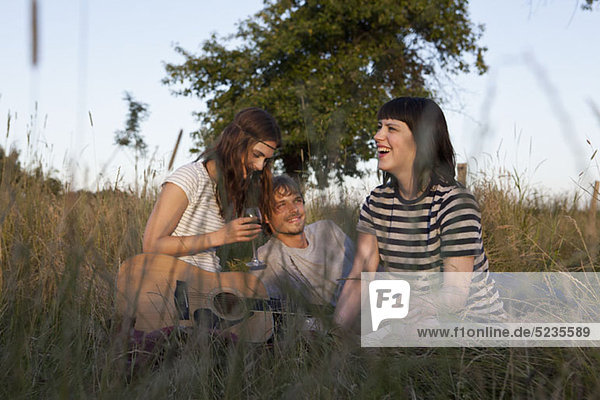 Three friends enjoying sitting in secluded field with wine and guitar