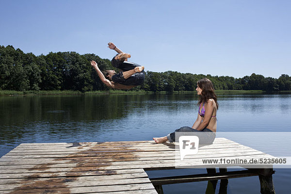 Der Typ springt mit ausgestreckten Armen in den See  während das Mädchen am Steg zuschaut.