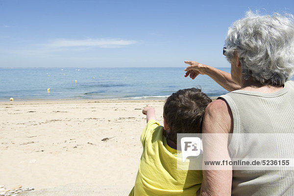 Großmutter und Enkel sitzen zusammen am Strand und schauen aufs Meer.