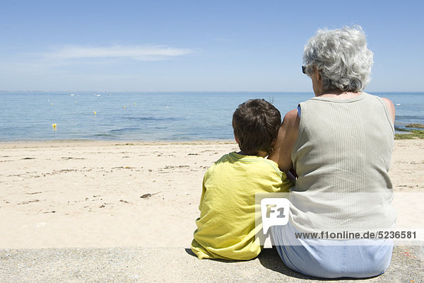 Großmutter und Enkel sitzen zusammen am Strand und schauen aufs Meer.