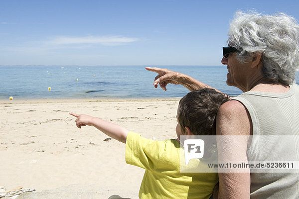 Großmutter und Enkel sitzen zusammen am Strand und zeigen auf das Meer.