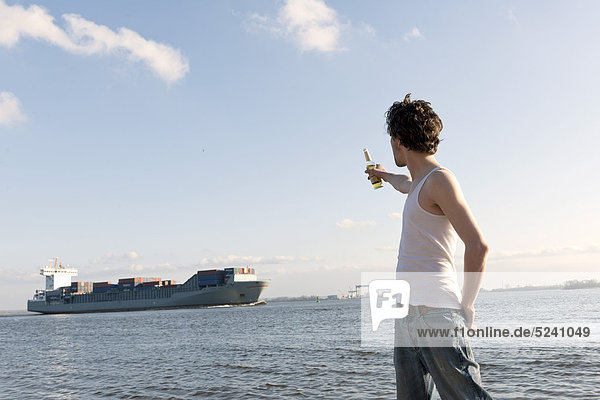 Mann mit Bierflasche am Elbufer und Containerschiff im Hintergrund