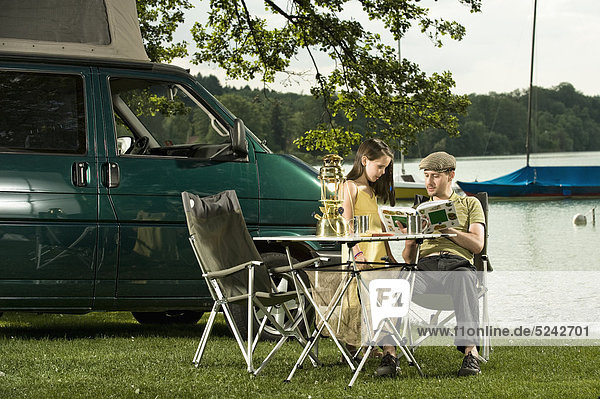 Germany  Bavaria  Woerthsee  Father and daughter reading book near lakeshore while camping