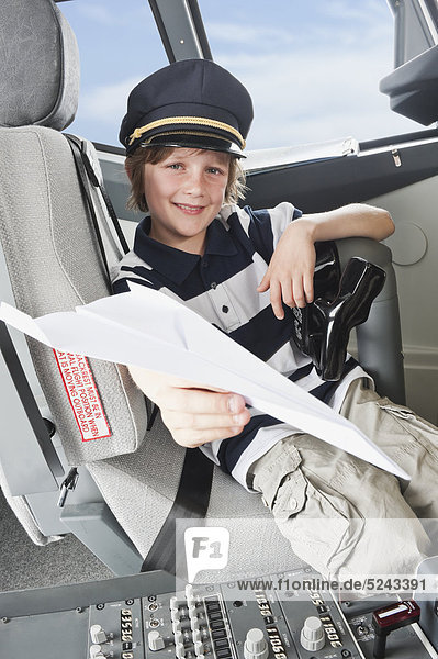 Boy wearing captain's hat and holding paper plane in airplane cockpit