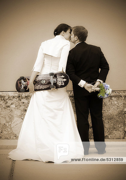 Kissing wedding couple seen from behind  the bride is holding a skateboard  the groom is wearing handcuffs and holding a bridal bouquet