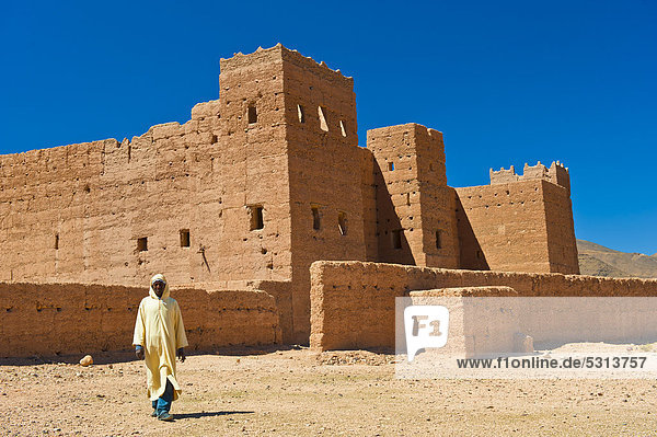 Berber man wearing a traditional djellabah walking in front of Taouirt Kasbah  mud fortress  mud brick building of the Berber tribes  Tighremt  Draa Valley  Southern Morocco  Morocco  Africa