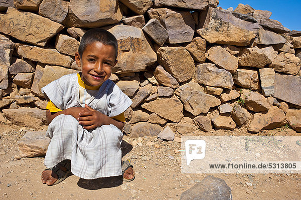 Small friendly boy wearing a traditional djellabah garment squatting in front of a stone wall  Agadir Tasguent  Anti-Atlas Mountains  southern Morocco  Morocco  Africa