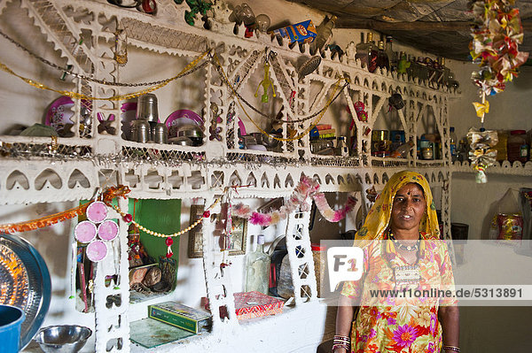Eine Inderin im traditionellen Sari steht in ihrer Küche an einem kunstvoll verzierten Regal für Haushaltsgeräte  Wüste Thar  Rajasthan  Indien  Asien