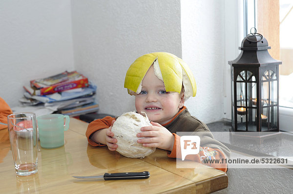 Junge  2 Jahre  sitzt mit Pomeloschale auf dem Kopf am Tisch