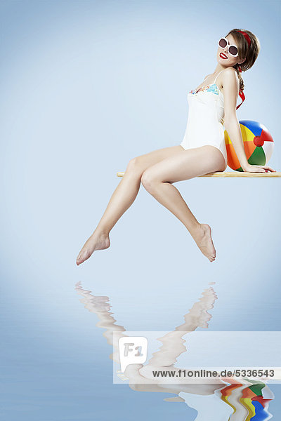 Junge Frau in einem hellen Badeanzug und Sonnenbrille sitzend auf einem Sprungbrett  hinter ihr ein Wasserball  Pin-up