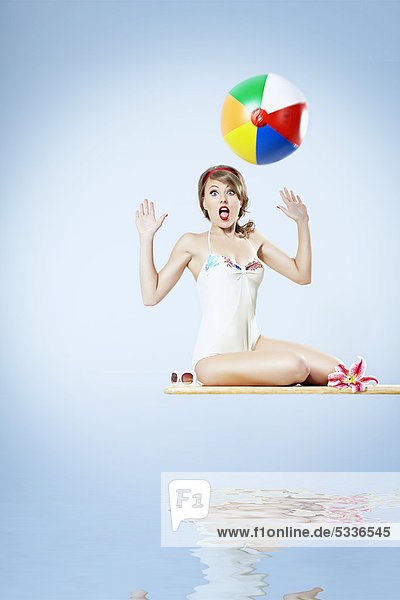 Junge Frau in einem hellen Badeanzug sitzt auf einem Sprungbrett und versucht einen Wasserball zu fangen  Pin-up