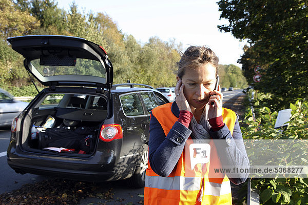 Car breakdown  female driver has stopped on the hard shoulder of a country road  wearing a reflective vest  makes call for help on her mobile phone