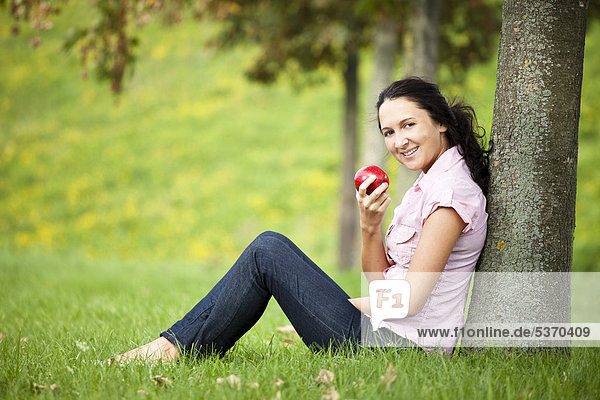Junge Frau lehnt an einem Baum  ißt einen roten Apfel