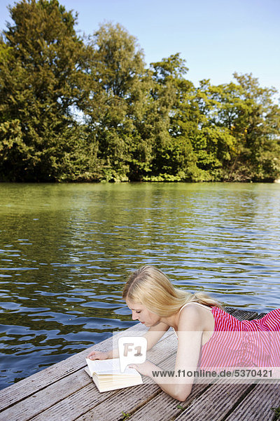 Junge Frau liegt ein Buch lesend auf einem Steg am Wasser