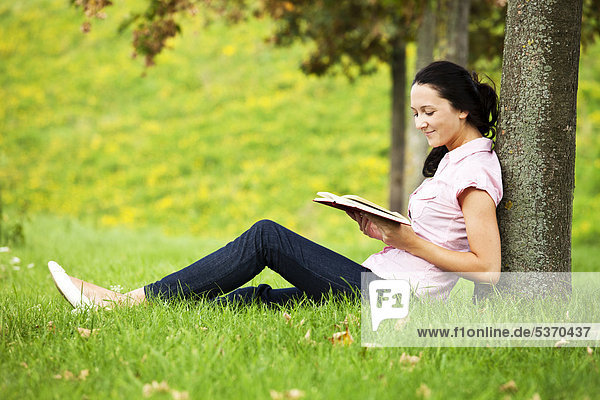 Junge Frau lehnt an einem Baum  liest ein Buch