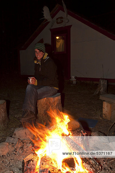 Young woman sitting at a camp fire  bonfire  holding a cup  illuminated wall tent  cabin with moose antlers behind  Yukon Territory  Canada