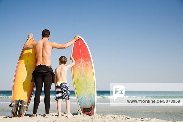 Vater und Sohn mit Surfbrettern  die aufs Meer hinausblicken.