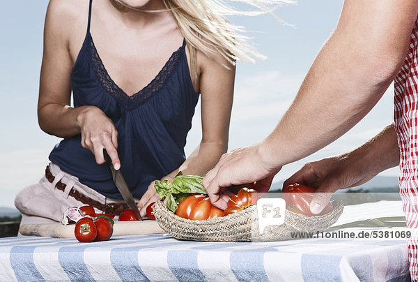 Italien  Toskana  Magliano  Junger Mann steht am Tisch und Frau schneidet Tomaten  lächelnd