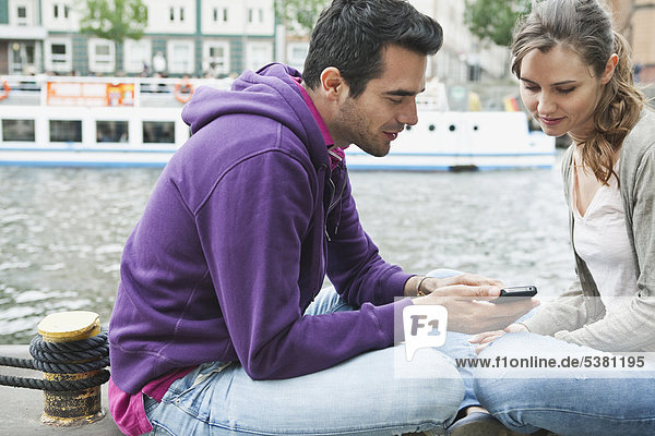 Deutschland  Berlin  Paar mit Handy am Flussufer