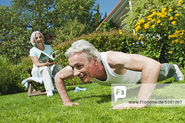 Mann trainiert und Frau liest Zeitschrift im Garten  lächelnd