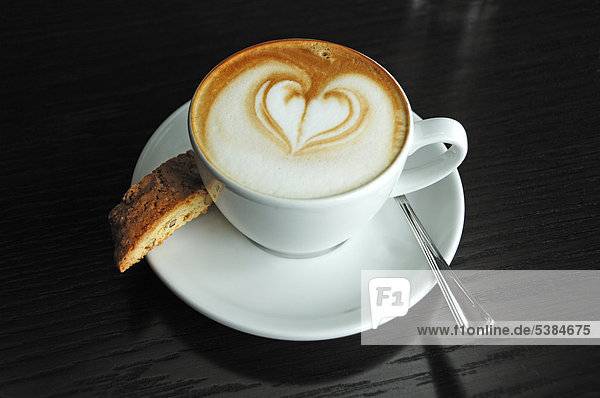 Tasse Cappuccino mit einem Herz verziert und einem Biscotti auf einem schwarzen Tisch