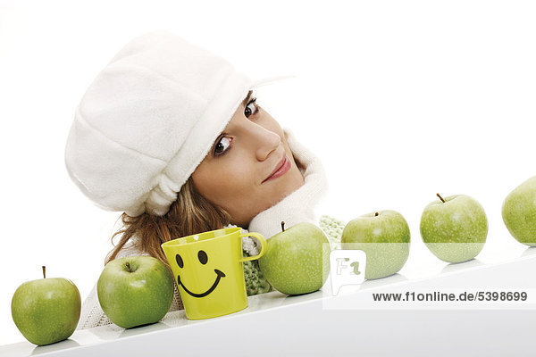 Young woman wearing a scarf and a cap  behind a row of green apples and a smiley mug