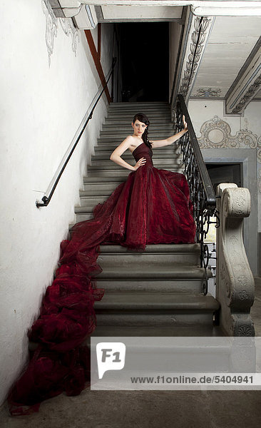 Junge Frau mit rotem Kleid und langer Schleppe sitzt auf Steintreppe in Treppenhaus