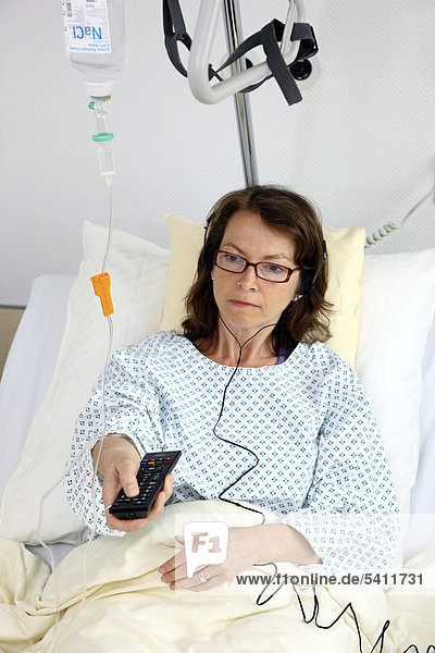 Patientin im Krankenbett  bedient mit der Fernbedienung einen Fernseher  hört den Ton über Ohrhörer  Krankenhaus
