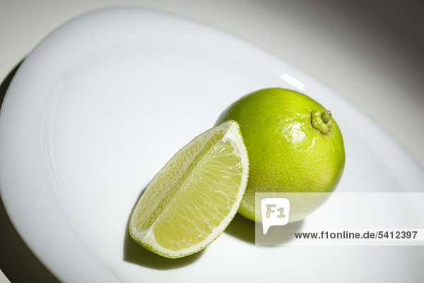 Limette (Citrus latifolia) und Limettenstück auf einem weißen Teller