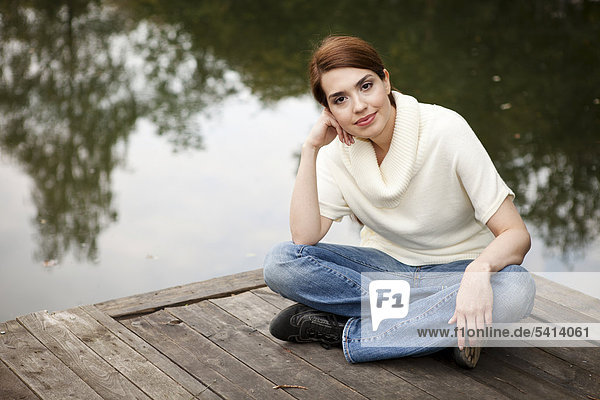 Junge Frau sitzt freundlich lächelnd auf einem Steg am See
