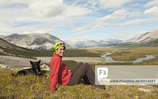Junge Frau sitzt  ruht aus  genießt Aussicht  Panorama  ein Alaskan Husky Schlittenhund neben ihr  Wind River und Mackenzie Mountains hinten  Peel Watershed  Yukon Territory  Kanada