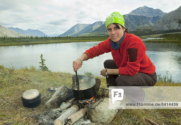 Young woman cooking on a camp fire  steering in a steaming pot  camping  Mackenzie Mountains behind  Wind River  Peel Watershed  Yukon Territory  Canada