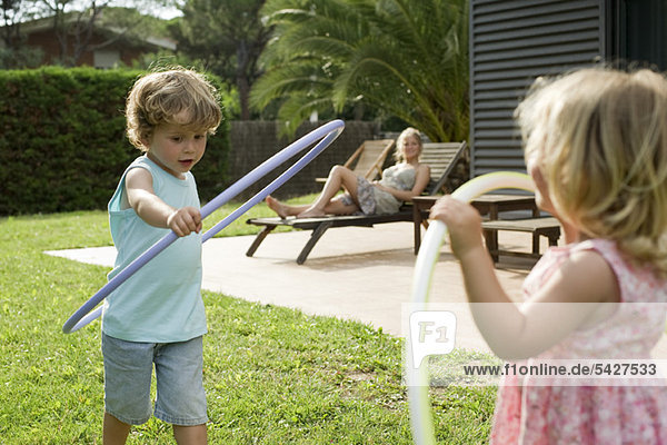 Kinder spielen mit Plastikbändern  während die Mutter im Hintergrund zuschaut.