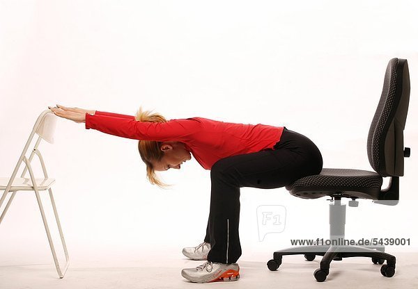 fit im Büro - junge Frau sitzt auf Bürostuhl und macht Gymnastik - Oberkörper auf Oberschenkel und Arme nach vorne auf Stuhl auflegen - Beweglichkeit