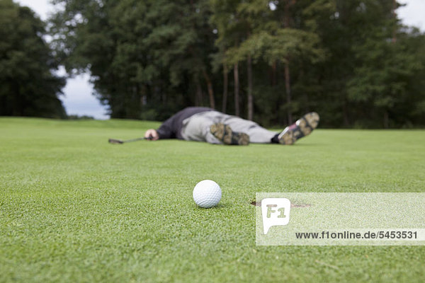 Ein verzweifelter Golfer  der auf dem Putting Green mit Ball am Lochrand liegt