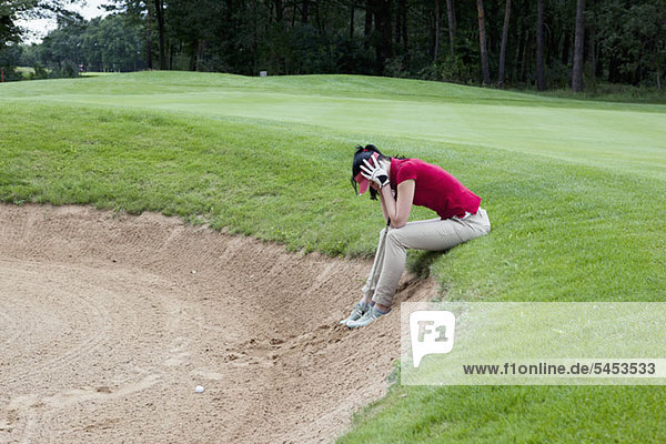 Eine Golferin sitzt am Rande einer Sandfalle  Kopf in den Händen.