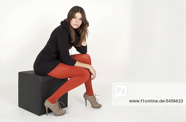 Young woman wearing a long black jumper and red tights  sitting on black cube