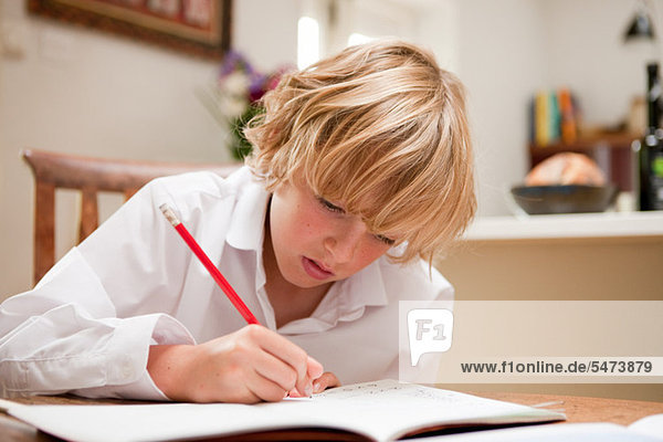 Young boy at home writing in school text book at the dining table