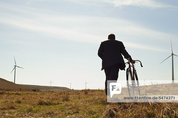 Man pulling bicycle uphill towards windfarm