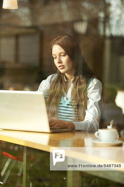 Studentin mit Laptop sitzt in Cafe  durch Fensterscheibe gesehen