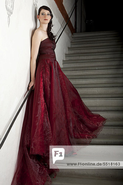 Junge Frau mit rotem Kleid und langer Schleppe steht auf Steintreppe in Treppenhaus