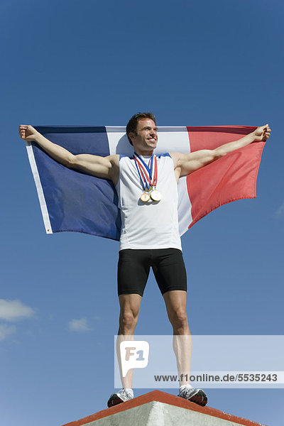 Männlicher Athlet wird auf dem Podium geehrt und hält die französische Flagge hoch.