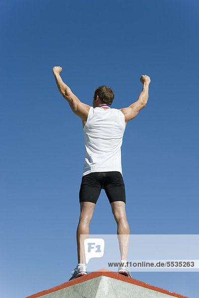 Male athlete standing on winner's podium with arms raised in victory  rear view