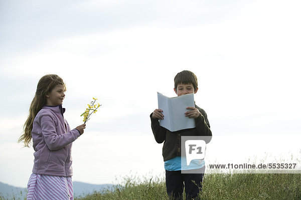Kinder im Feld  Junge liest Buch  während Mädchen Wildblumen pflückt.