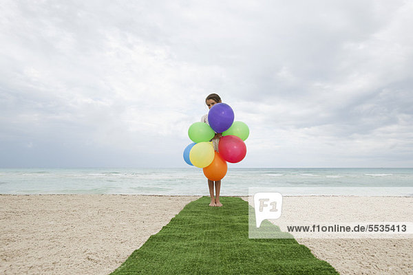 Mädchen steht auf Kunstrasen am Strand  Gesicht versteckt hinter Luftballons