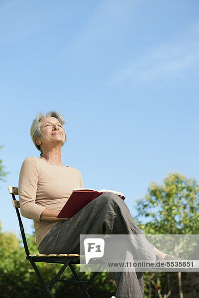 Seniorin im Freien sitzend mit Buch  Sonnenwärme genießend