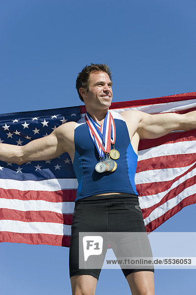 Männlicher Athlet auf dem Siegerpodest  der die amerikanische Flagge hochhält.