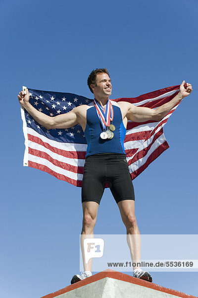 Männlicher Athlet auf dem Siegerpodest  der die amerikanische Flagge hochhält.
