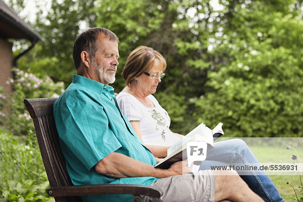 Seniorenpaar liest Buch im Garten  während es im Sessel sitzt.
